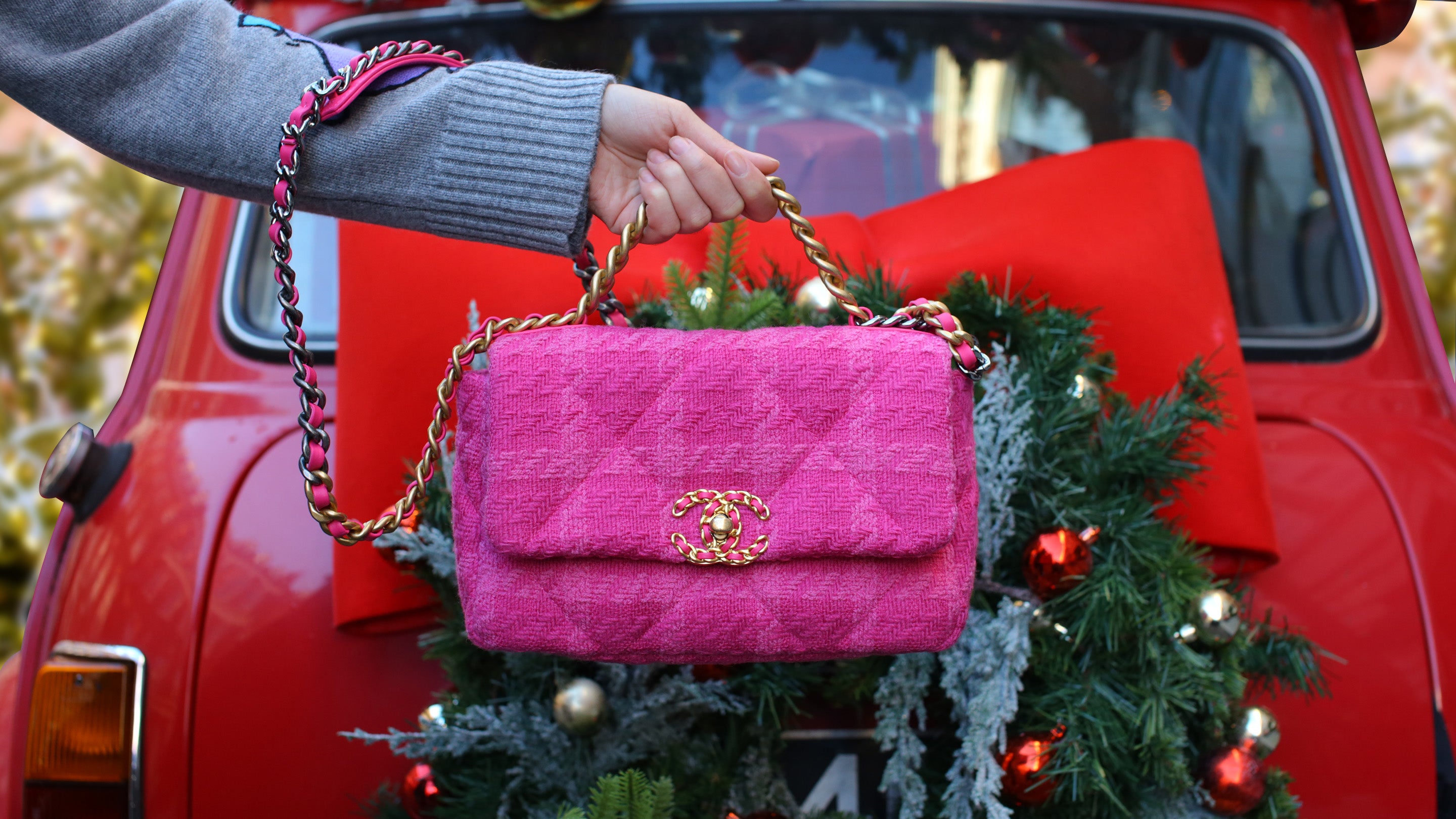 Pink quilted Chanel 19 Flap Bag with gold chain held by a person in front of a red car with Christmas decorations.