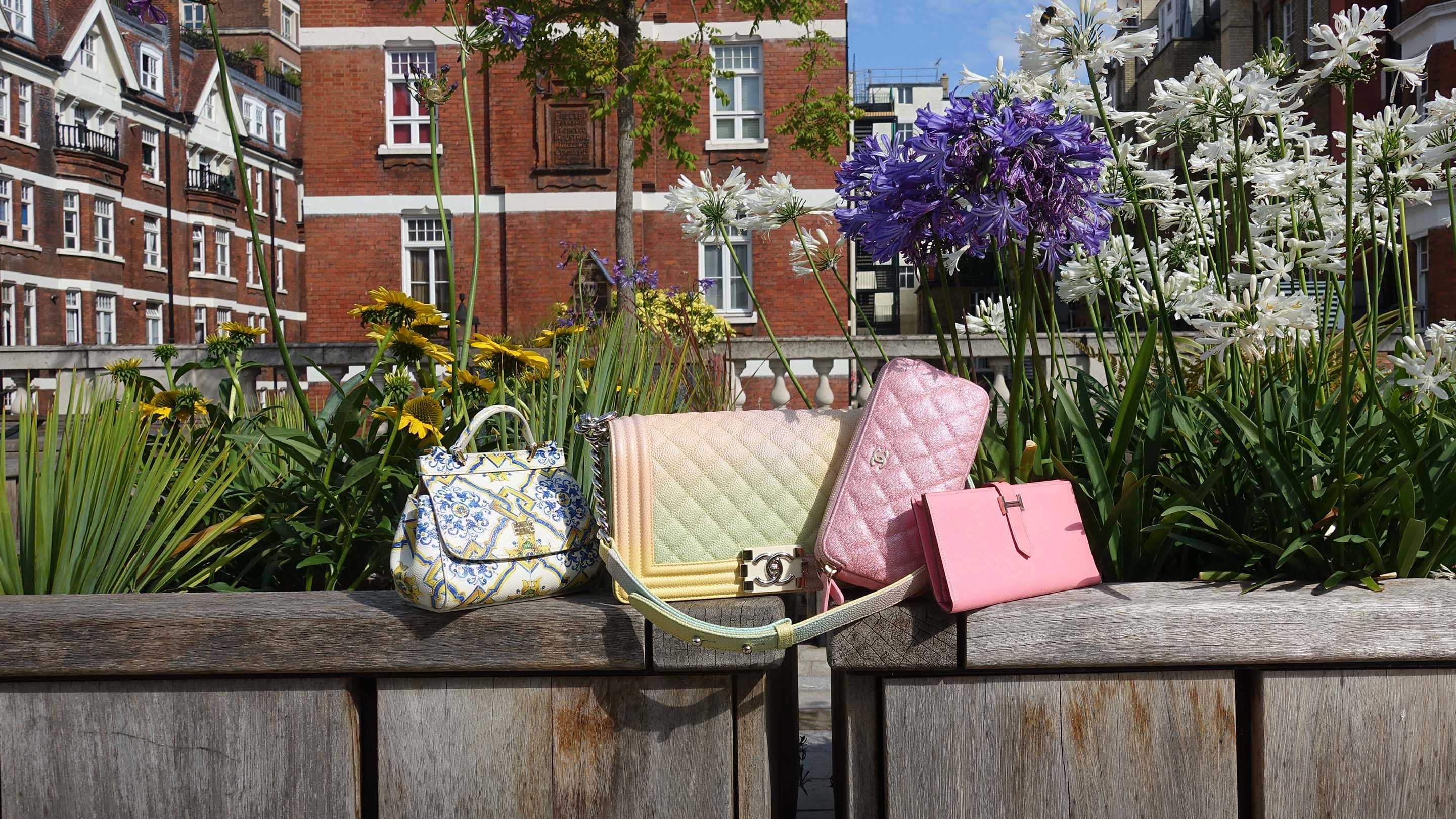 Preloved Chanel bags, Hermes wallet, and Dolce & Gabbana bag displayed on a wooden ledge with a garden and building in the background.