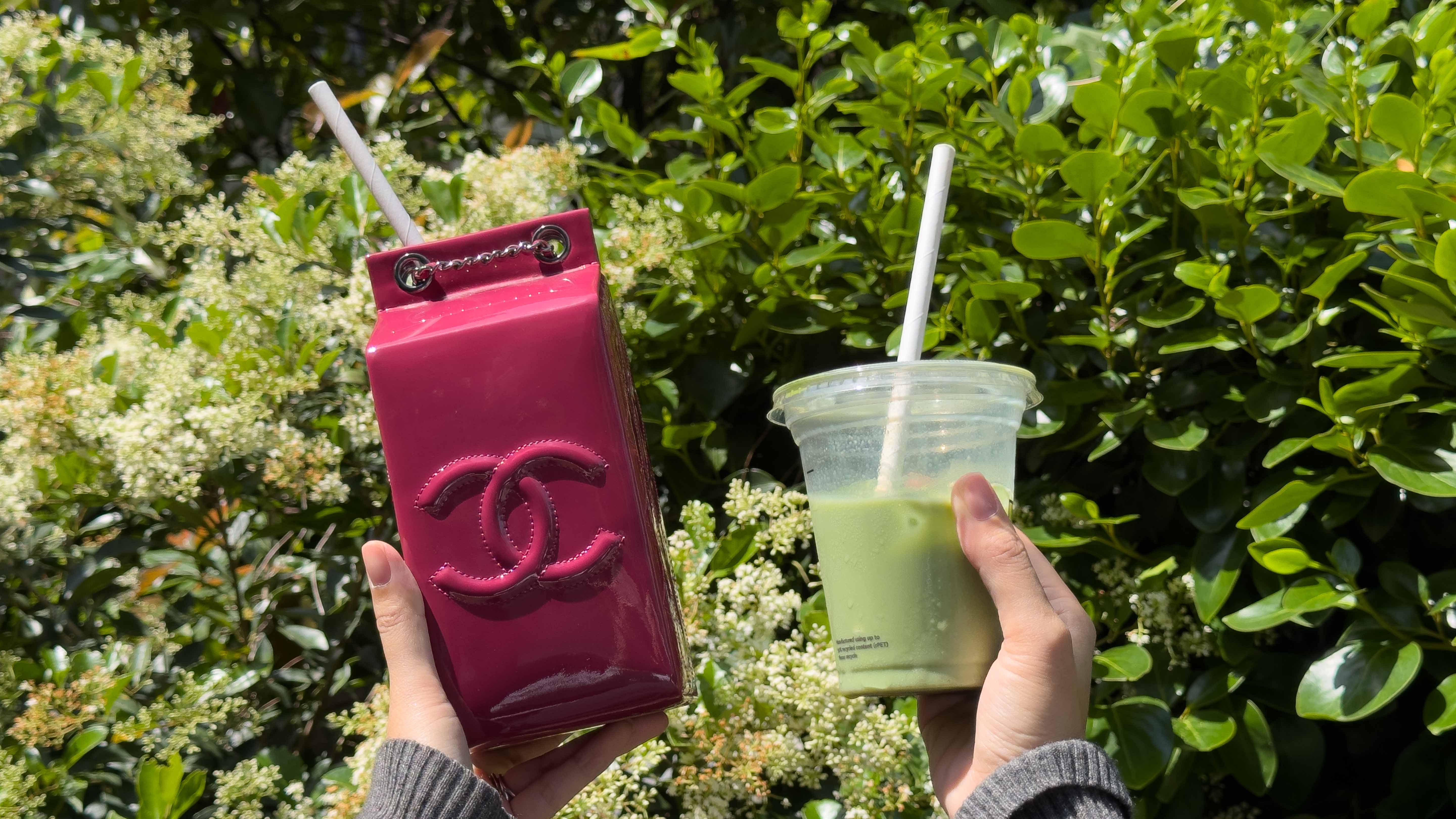 A person holding a rare preloved fuchsia Chanel milk carton handbag alongside an iced matcha latte against a backdrop of lush summer greenery.