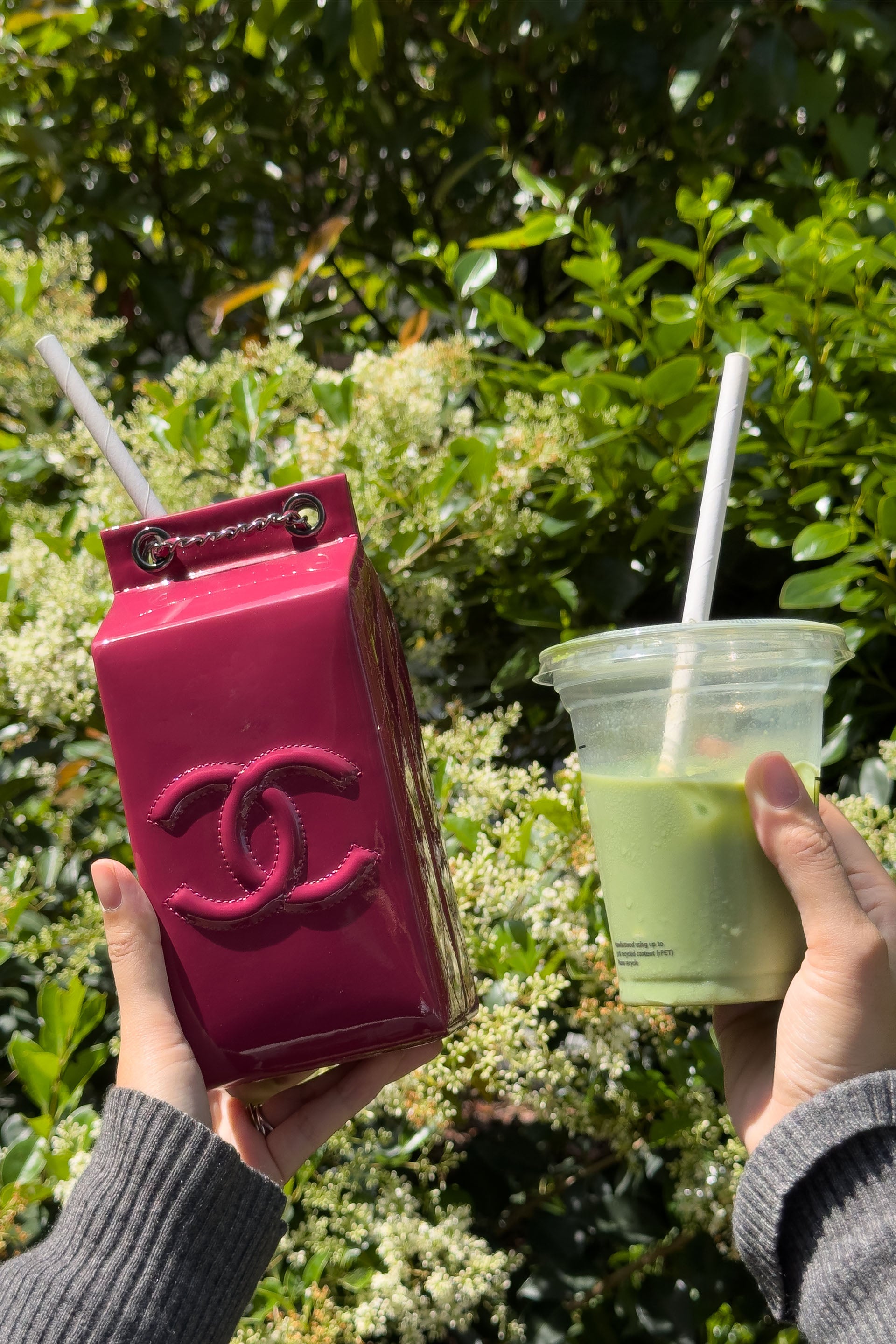 Two hands holding a pink Chanel milk carton bag and a green drink with a straw against a green leafy background.